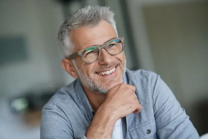 Silver haired man with wood grain glasses learning on his hand while smiling