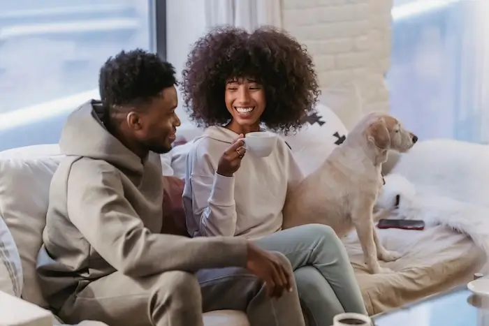 A couple sitting on a couch with a dog beside them, laughing together while the woman holds a coffee cup.