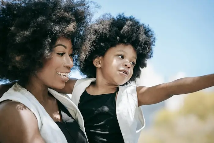 A joyful woman with curly hair smiling and looking at her child, who is playfully stretching out arms against a blue sky background.