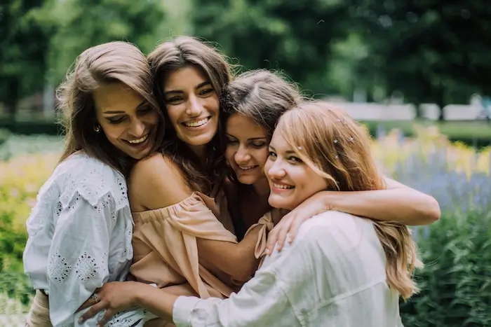 Four women hugging and smiling in a lush garden, with one looking directly at the camera.