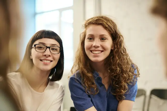 Two women, one with curly red hair and the other with straight black hair and glasses, smiling in an office environment.