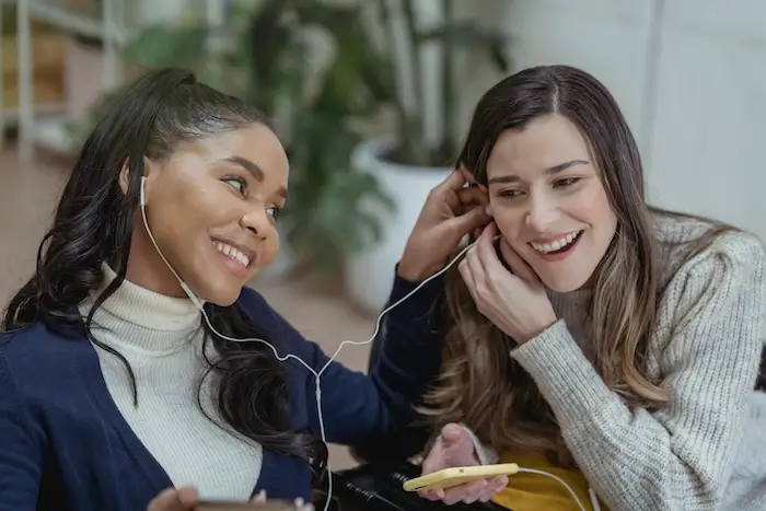 Two women, one with long curly hair and headphones, sharing a joyous moment with a smartphone and earbuds indoors.