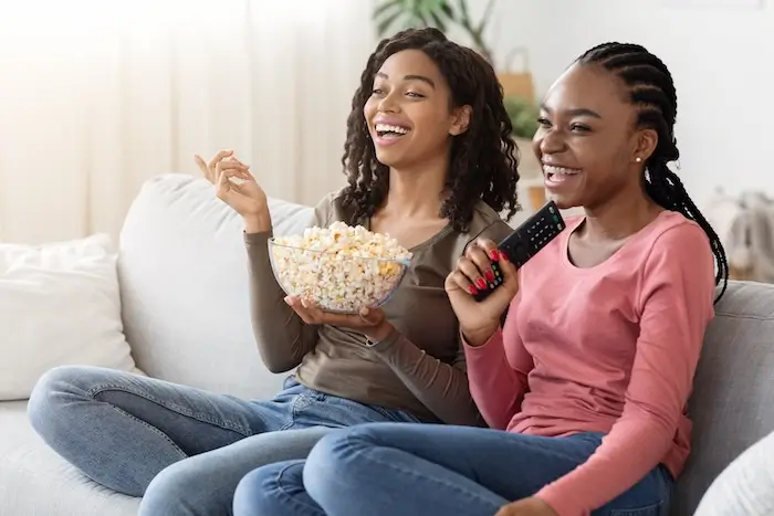 Two women laughing while sitting on the couch watching television and eating popcorn