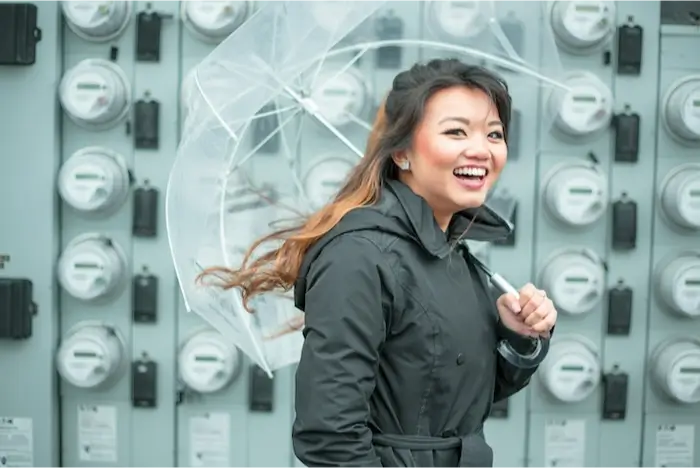 A cheerful woman with an open clear umbrella smiling broadly, standing in front of a bank of electricity meters.
