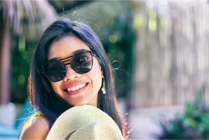 A happy woman in sunglasses and a hat, with a bright smile, looking over her shoulder in a sunny, tropical setting.