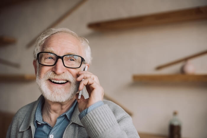 An older man with black frame glasses smiling while talking on his cellphone.
